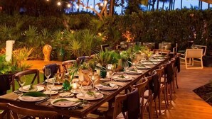 An outdoor dining area with tables set for a meal, surrounded by greenery and string lights.