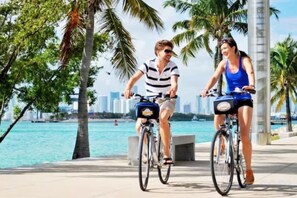 Two people riding bicycles on a paved path with palm trees and a body of water in the background. - Casagrandesouthbeach by AmericanVacationLiving 4Rm (Miami Beach)