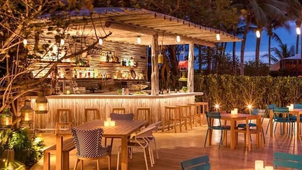 An outdoor bar area with wooden tables and chairs, surrounded by greenery and palm trees.