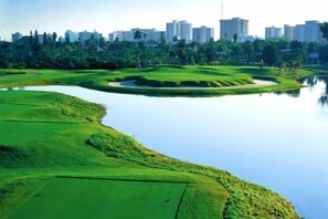 A golf course with a water body, surrounded by greenery and a city skyline in the background. - Casagrandesouthbeach by AmericanVacationLiving 4Rm (Miami Beach)