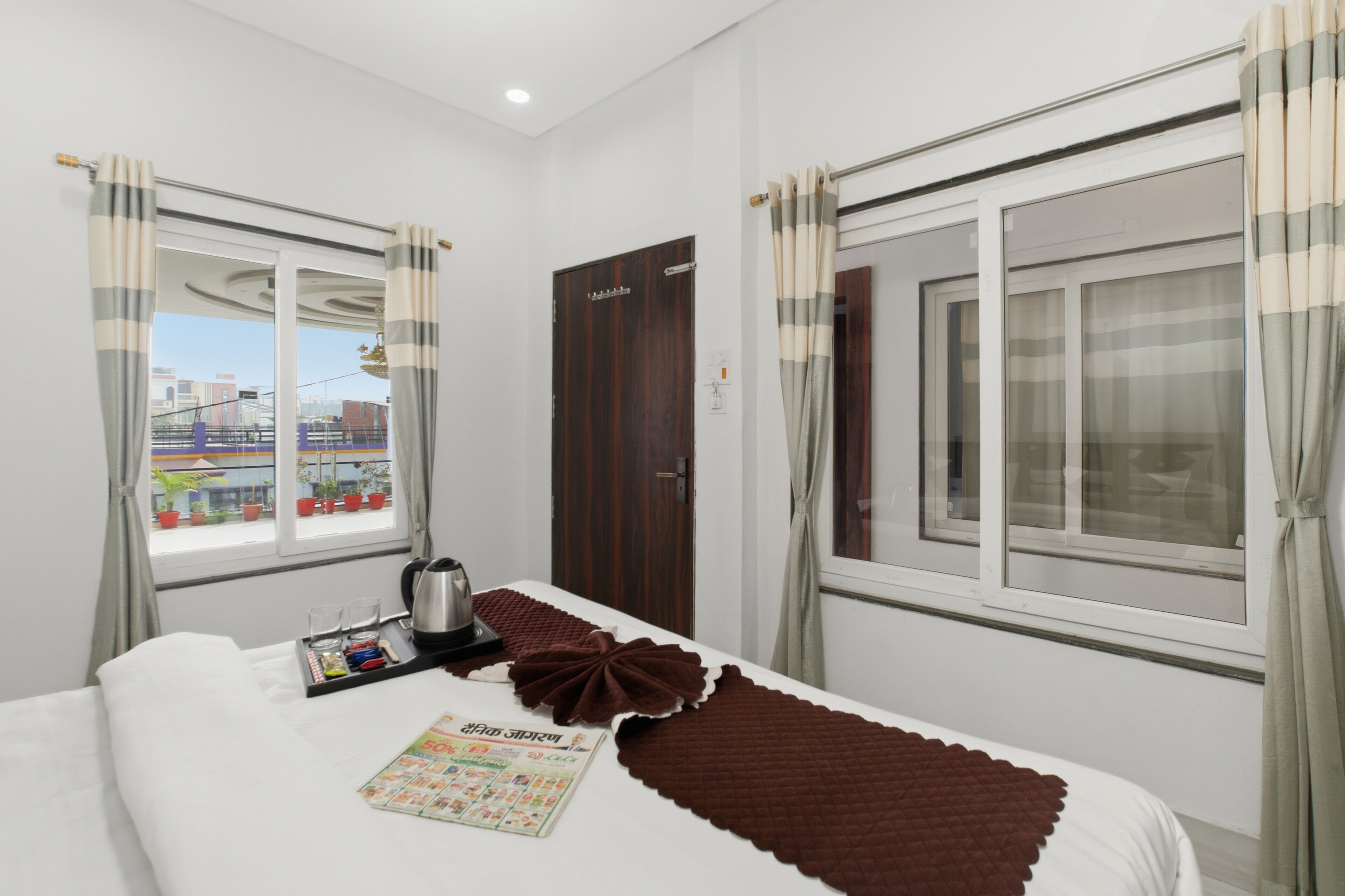 A hotel room with a bed, a window with curtains, a door, and a view of the cityscape.