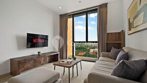 A modern living room with a flat-screen TV, a sofa, a coffee table, and a view of a residential area through large windows. - Oswin by Kozystay - Cilandak (Jakarta)