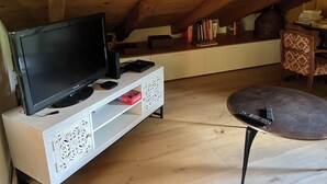 Una habitación con televisión sobre un mueble blanco, una mesita pequeña y una silla con cojín estampado.