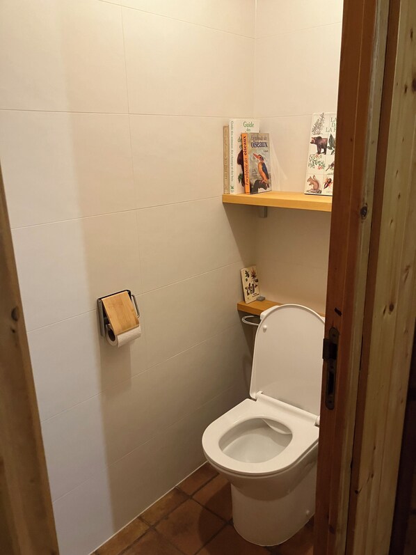 A small, clean bathroom with a toilet, a toilet paper holder, and a shelf with books and a small decorative item.
