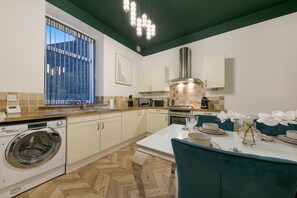 A modern kitchen with a built-in washing machine, a dining area with a table set for two, and a large window with a view of a building. - Rosemount Place Apartment - Grampian Lettings (Aberdeen)