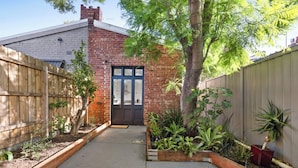 A narrow concrete pathway leading to a brick house with a dark front door, surrounded by a wooden fence and various plants. - Prime home near train, trams, cafes, schools, Yarra River & city attractions (Richmond)