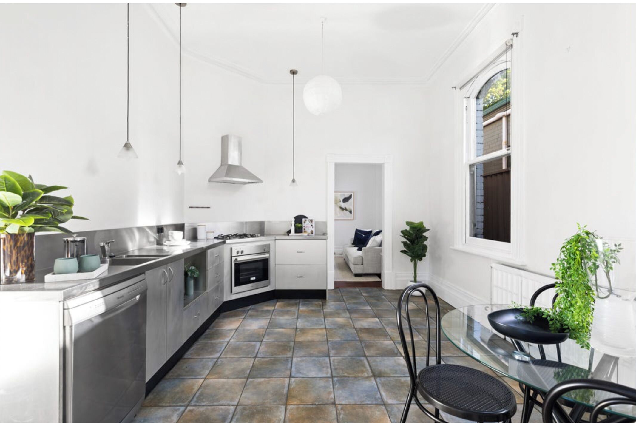A modern kitchen with stainless steel appliances, a dining area with a glass table, and a view of the garden through a window.