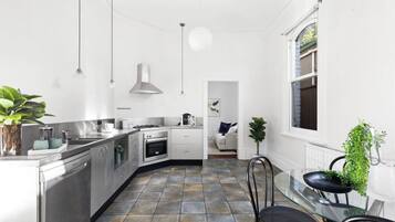 A modern kitchen with stainless steel appliances, a dining area with a glass table, and a view of the garden through a window.