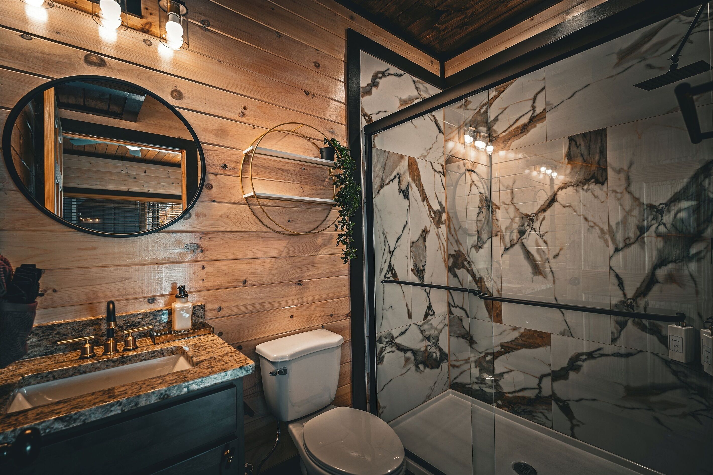 A modern bathroom with wooden paneling, a marble shower wall, a round mirror, and a granite countertop.