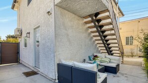 Uma varanda coberta ao ar livre com um sofá e almofadas, uma escada e uma porta que dá para um edifício.