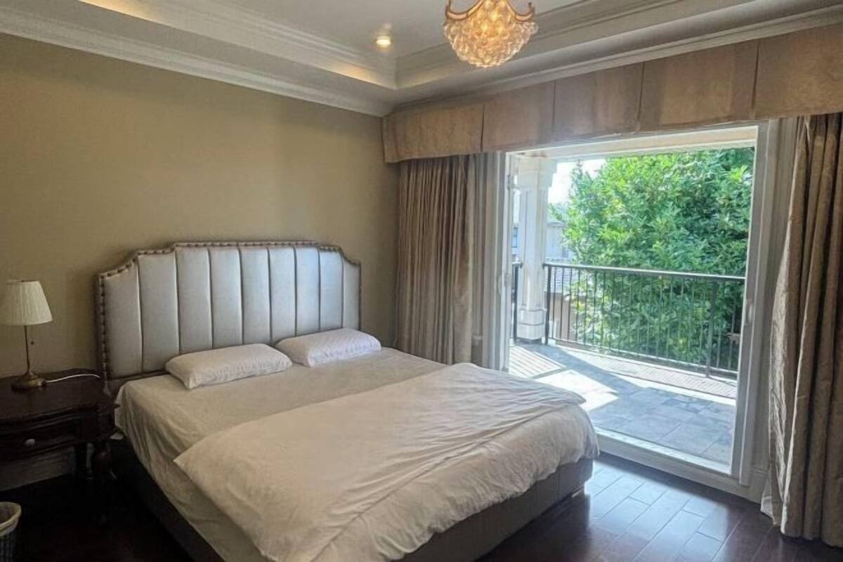 Habitación de hotel con una cama grande, una mesita de noche, una lámpara y un balcón con vistas a zonas verdes.
