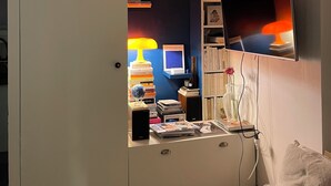 Huoneessa on valkoinen vaatekaappi, työpöytä, jossa on lamppu, kirjoja, televisio ja pehmeällä tyynyllä varustettu sohva.