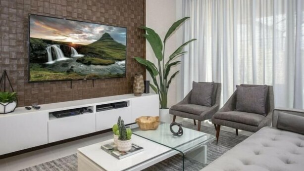 Ein modernes Wohnzimmer mit einem Flachbildfernseher an einer Ziegelwand, zwei grauen Sesseln, einem Glastisch und einer großen Pflanze.