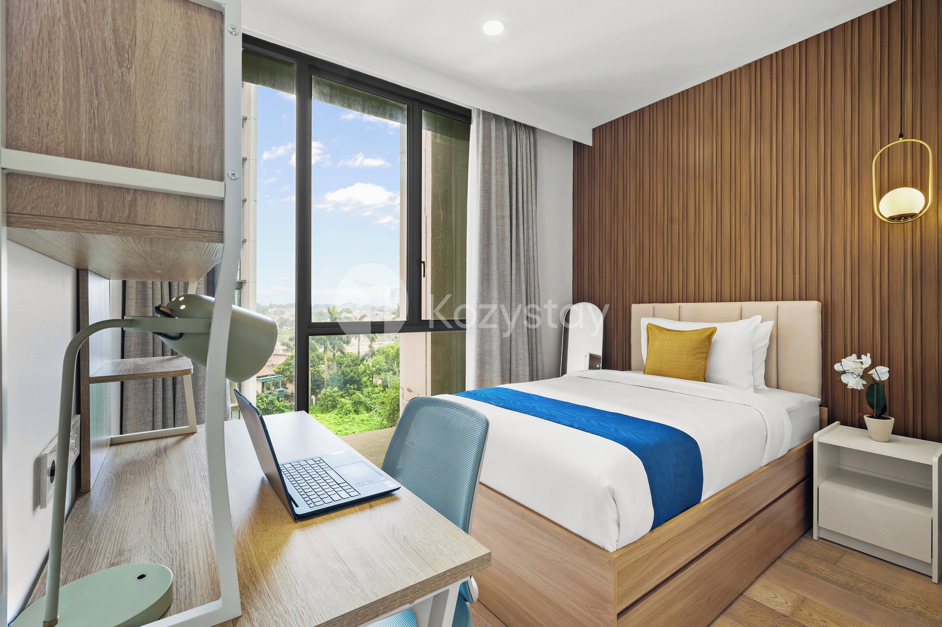 A modern hotel room with a wooden wall, a large bed with a blue and white bedspread, a desk with a laptop, and a view of greenery through the window.