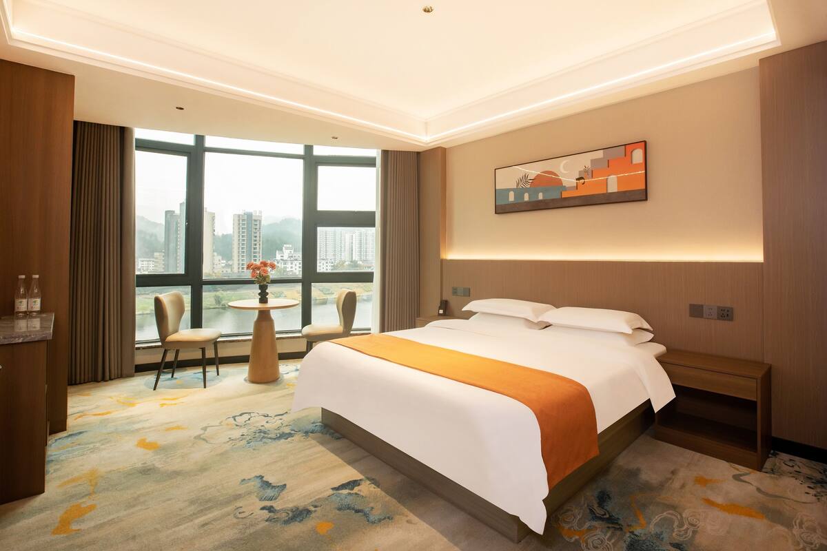 Habitación de hotel con una cama grande, un escritorio con silla, una mesita con un florero, y vistas a edificios a través de amplias ventanas.