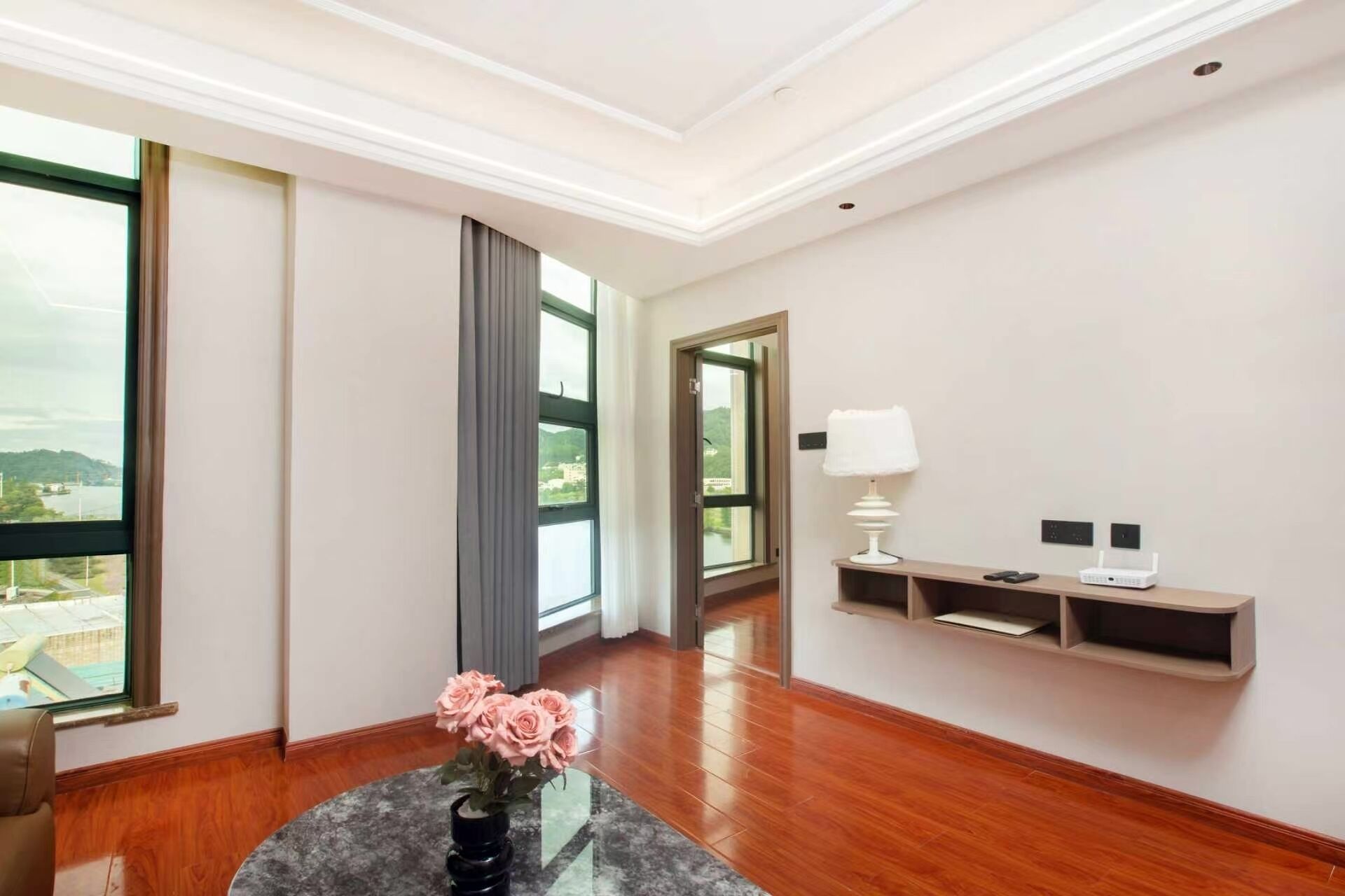 Une chambre d’hôtel avec un plancher en bois, une table ronde ornée d’un vase de fleurs, une fenêtre donnant sur un paysage verdoyant, une porte et une étagère murale avec une lampe et un routeur.