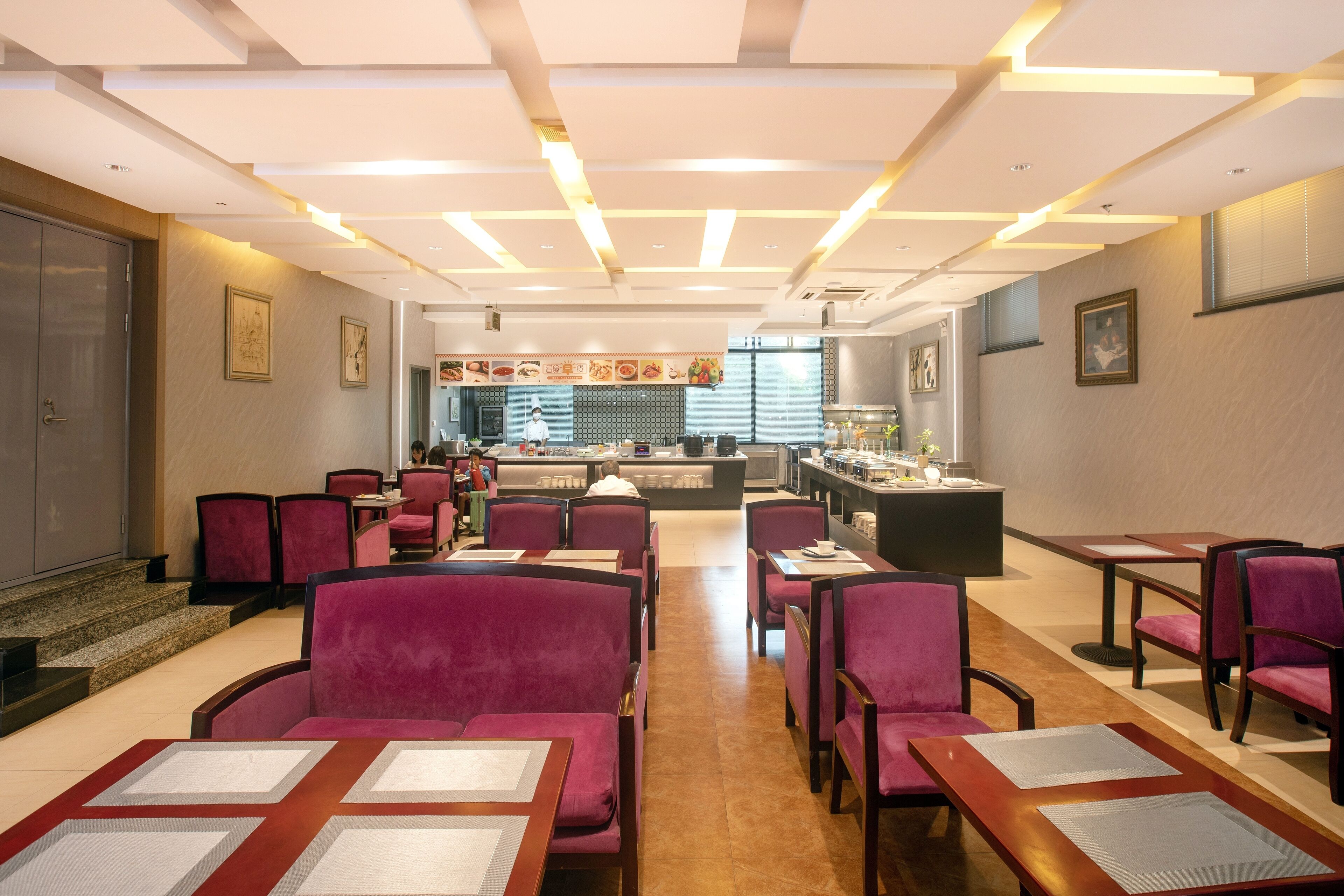 A modern restaurant with red velvet chairs and wooden tables, a kitchen in the background, and framed pictures on the walls.