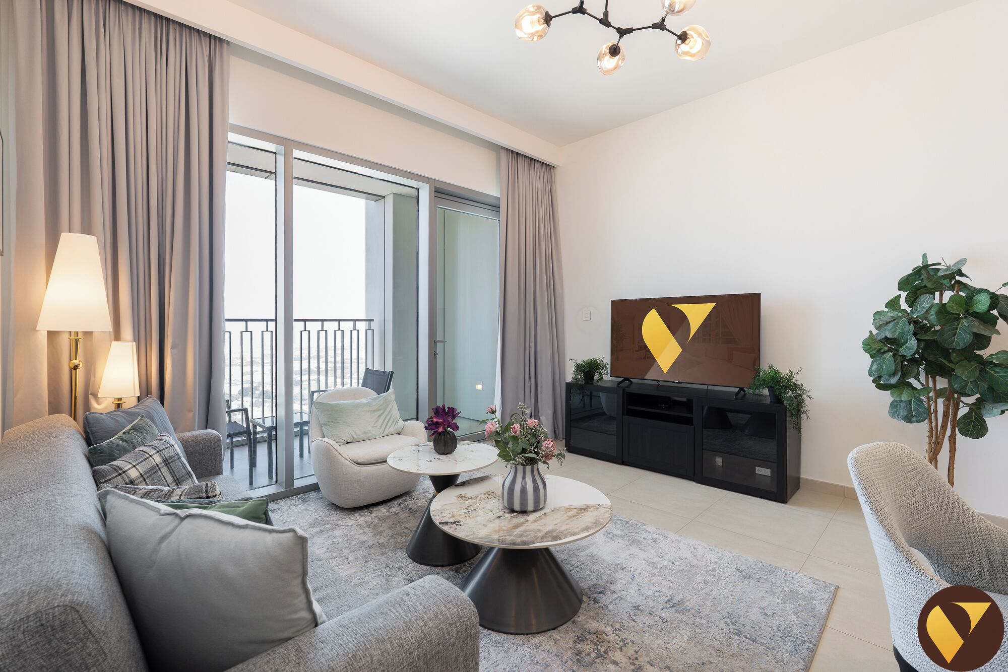 A modern living room with a sofa, armchairs, a coffee table, and a TV. There is a balcony with curtains and a plant.