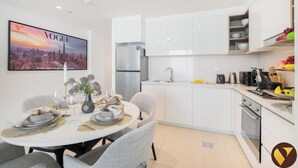 A modern kitchen with white cabinetry, a stainless steel refrigerator, and a dining area with a table set for four. - Vogue | Downtown with Direct Access to Dubai Mall (Dubai)