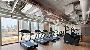 A modern gym with treadmills and elliptical machines, large windows offering city views, and exposed ductwork on the ceiling. - Vogue | Downtown with Direct Access to Dubai Mall (Dubai)