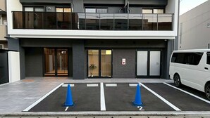 Un edificio moderno con una entrada de dos pisos, un estacionamiento con dos espacios señalizados y una camioneta blanca estacionada a la derecha.