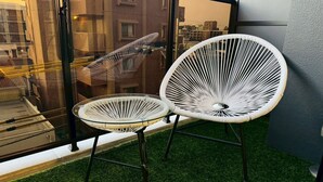 Dos sillas blancas de mimbre con patas metálicas en un balcón con césped artificial, con vista a un paisaje urbano.