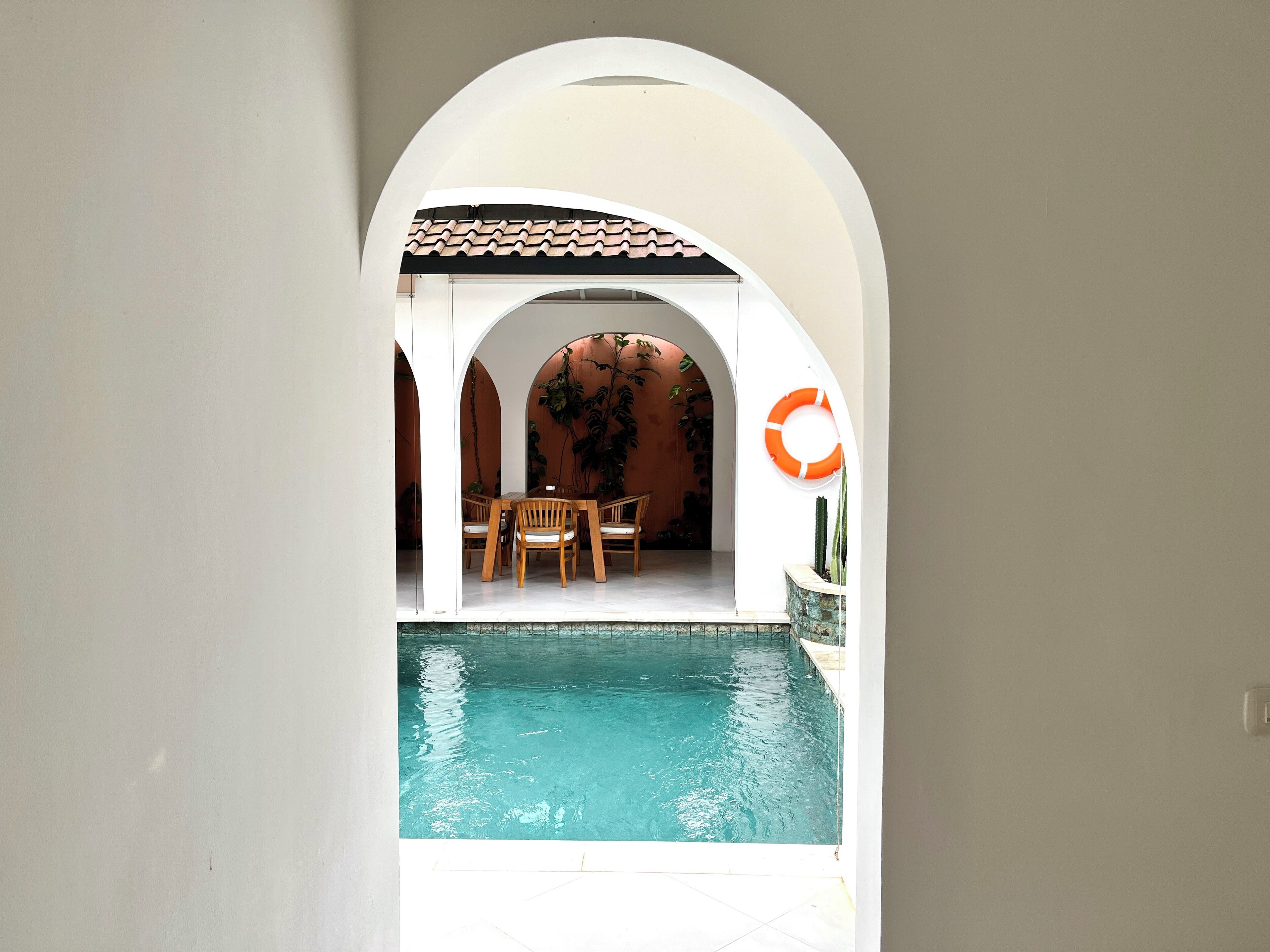 An indoor swimming pool with a tiled floor, surrounded by white walls and arches. There is a wooden table and chairs in the background, and a lifebuoy on the wall.