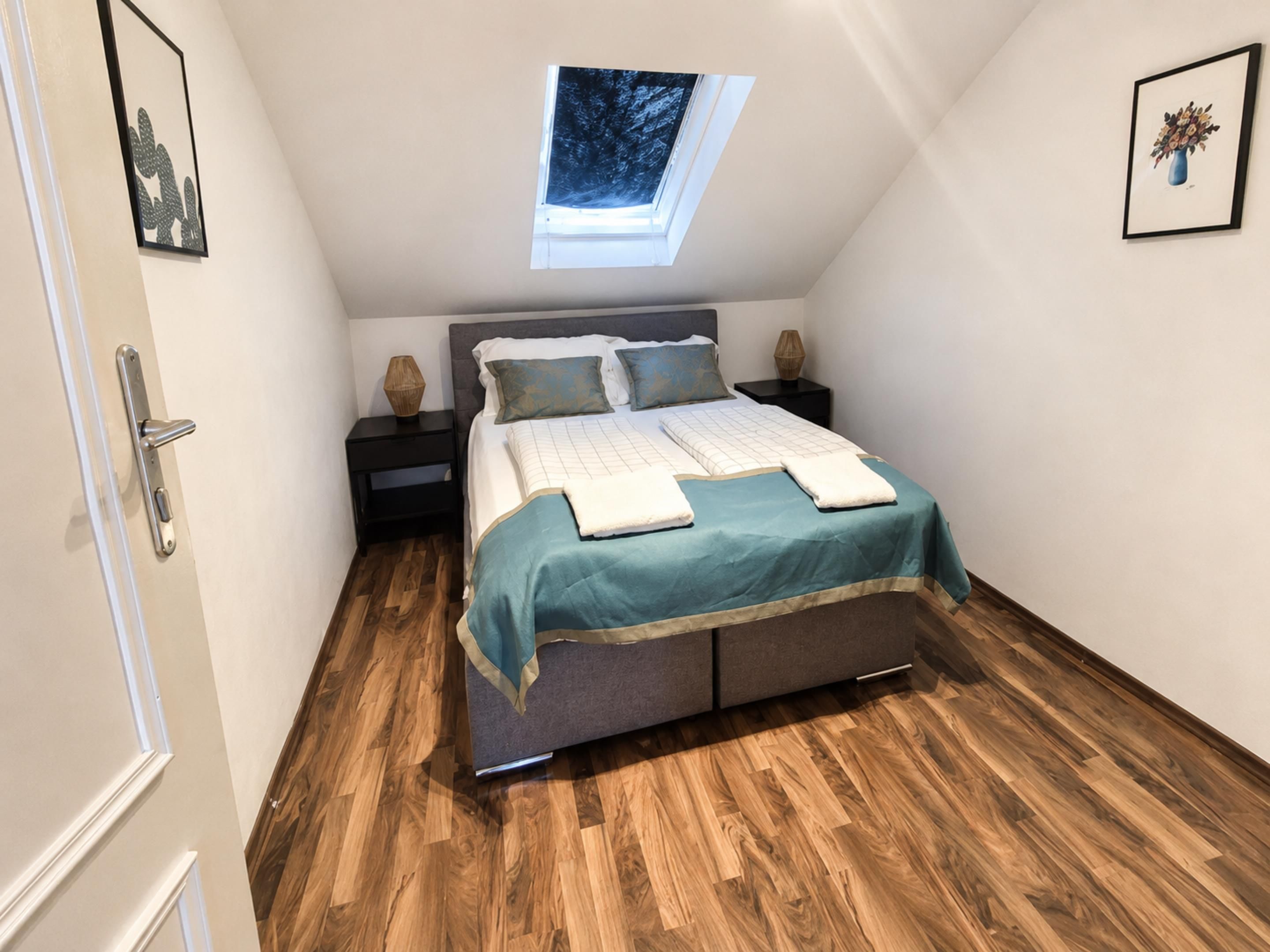 Un dormitorio con tragaluces, una cama con colcha color turquesa, dos mesitas de noche y suelo de madera.