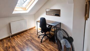 Una habitación con techo inclinado, tragaluz, radiador, escritorio con silla, espejo y ventilador.