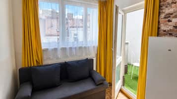 Una habitación con sofá, ventana con cortinas amarillas, puerta que da a un balcón y refrigerador blanco.