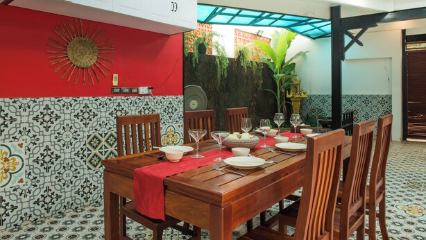 A dining area with a wooden table set for four, surrounded by chairs. There is a red tablecloth, a decorative wall with a sun motif, and a patterned tile floor. - Peaceful 4-Bedroom Villa with Private Pool (Siem Reap)