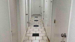A narrow hallway with white walls and a tiled floor, leading to a closed door at the end.