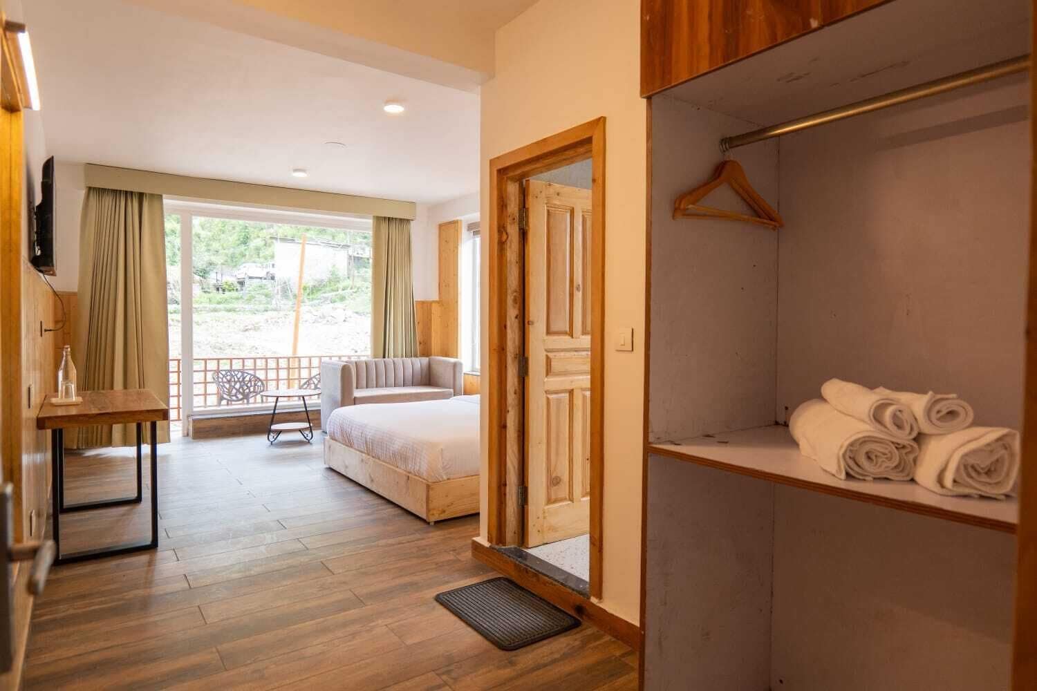 A hotel room with a bed, a seating area with a table and chairs, a wardrobe with hanging clothes, and a view of the outdoors.