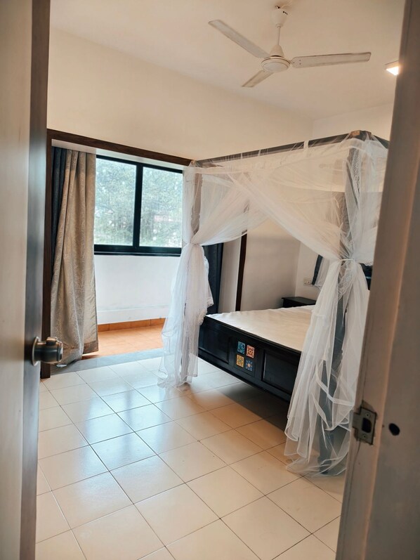 A bedroom with a bed under a canopy, a ceiling fan, a window with curtains, and a tiled floor. - Osho Apartments (Panchgani)