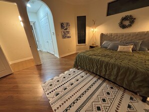 Ein Schlafzimmer mit einem Bett, einem gemusterten Teppich, einem Nachttisch, Wandbildern und einer Tür, die in einen anderen Raum führt.