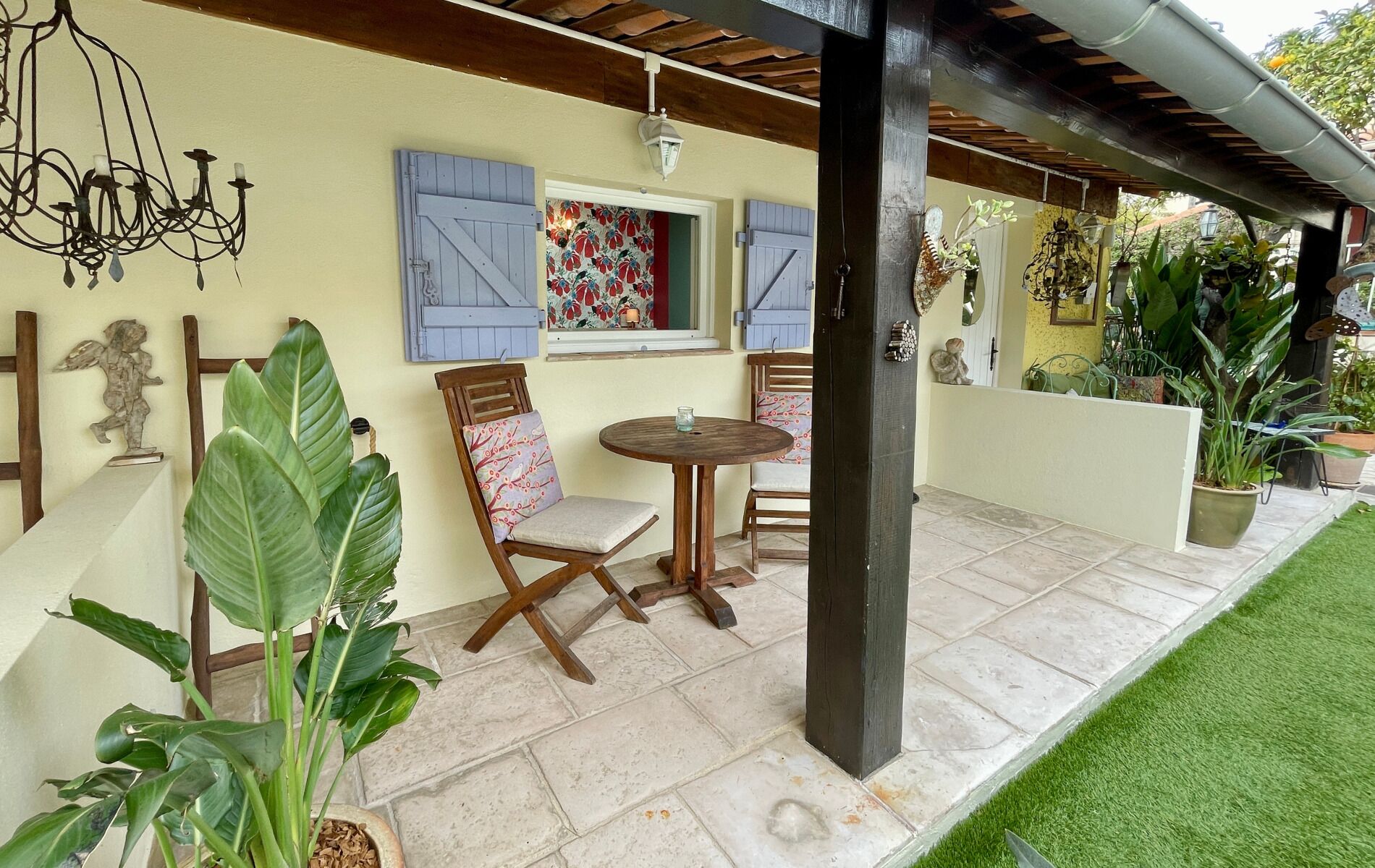 Un patio cubierto con una mesa y sillas, una pared decorativa con contraventanas y diversas plantas.