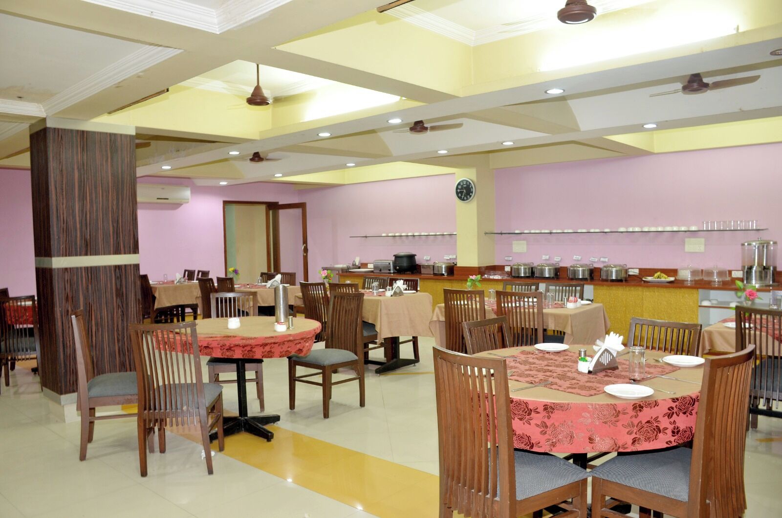 Un comedor con varias mesas y sillas, un reloj en la pared y una barra de buffet con diversos alimentos.