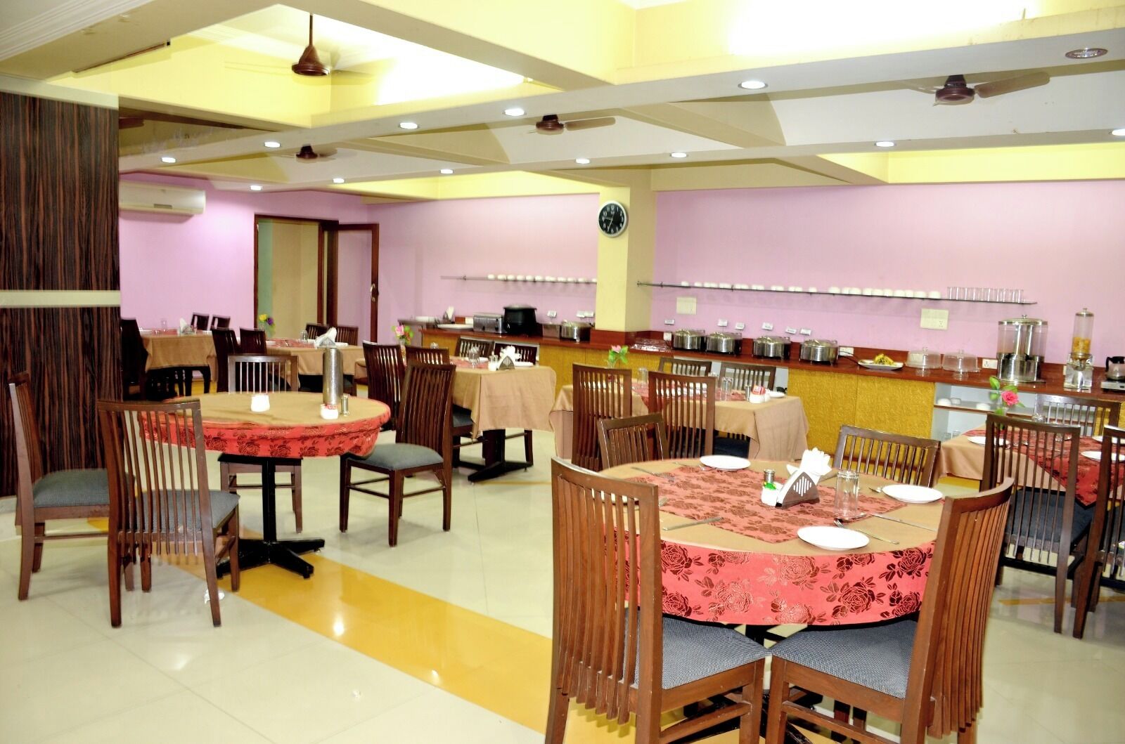 A dining area with round tables, chairs, and a buffet line with food containers.