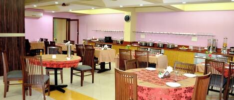 A dining area with round tables, chairs, and a buffet line with food containers.