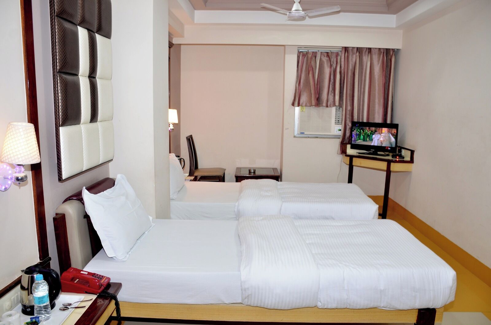 Habitación de hotel con dos camas, televisión, escritorio con lámpara, una silla y ventana con cortinas.