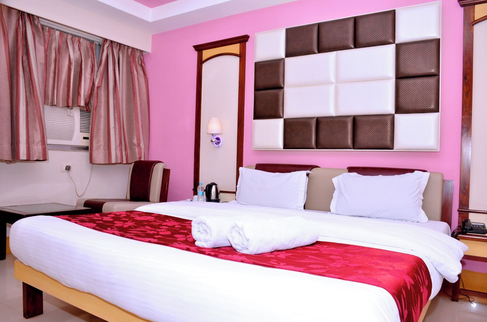 A hotel room with a large bed, a bedside table, a chair, an air conditioning unit, and a mirror.