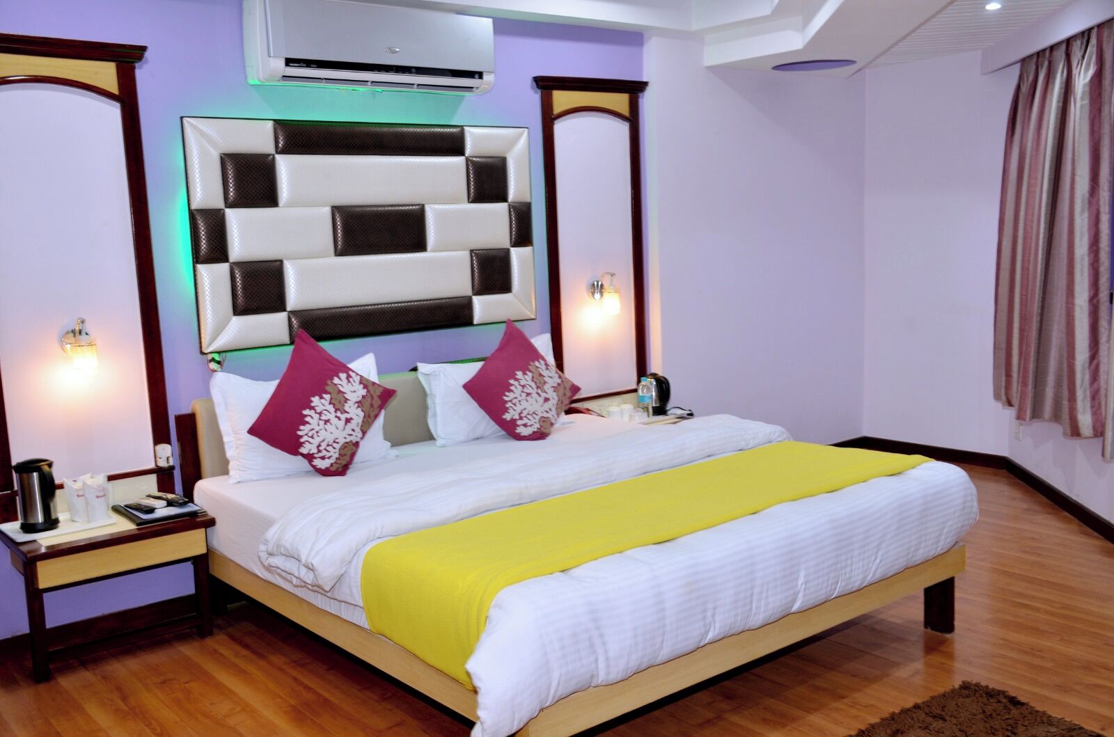 A hotel room with a large bed, a bedside table, an air conditioning unit, and a wall-mounted light fixture.