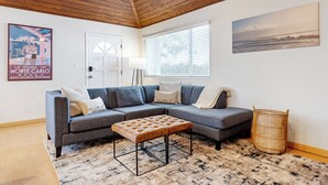 A modern living room with a grey sectional sofa, a tufted ottoman, a wooden ceiling, a beach-themed poster, a window with blinds, and a lamp. - Beach Home with Open Living & Walkability to Abbot Kinney | Venice Drift (Los Angeles)