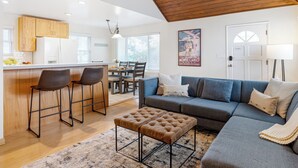 A modern living room with a grey sofa, a brown ottoman, and a kitchen area with wooden cabinets and a bar stool. - Beach Home with Open Living & Walkability to Abbot Kinney | Venice Drift (Los Angeles)