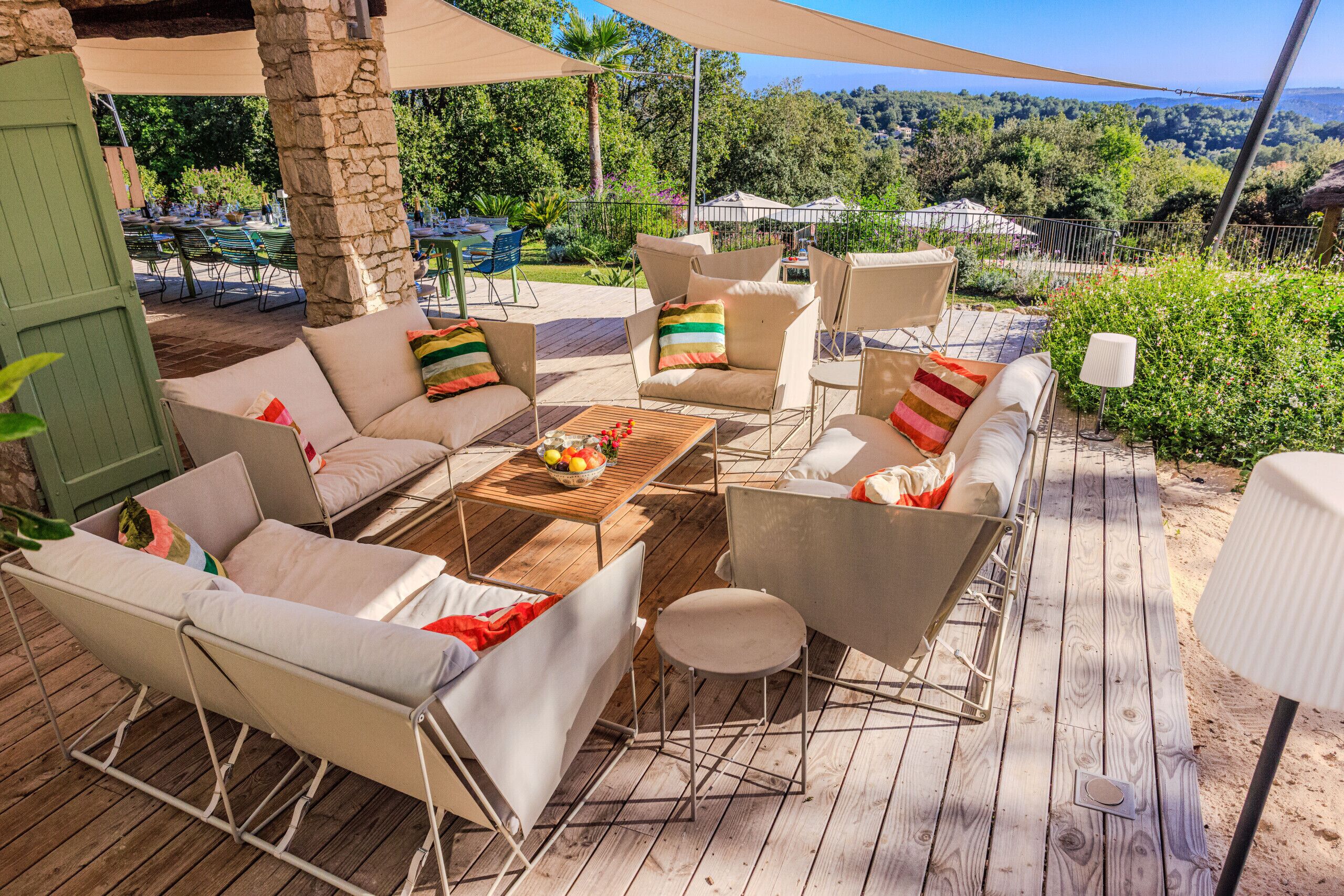 Une terrasse extérieure dotée d’une plateforme en bois, de chaises beiges avec des coussins, d’une table ronde sur laquelle se trouve un panier de fruits, et qui offre une vue sur un paysage luxuriant.