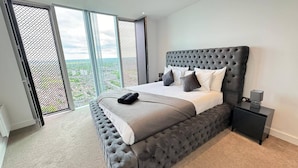 Un dormitorio moderno con una cama grande, cabecera tapizada en gris y vistas a la ciudad a través de una puerta corrediza de cristal.