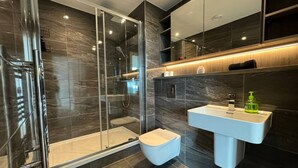 Un baño moderno con ducha envuelta en cristal, un lavabo blanco y paredes de azulejos oscuros.