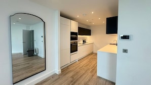 Una cocina moderna con armarios blancos, horno integrado y microondas. En la pared izquierda hay un espejo grande y, a la derecha, un pequeño rincón con fregadero.