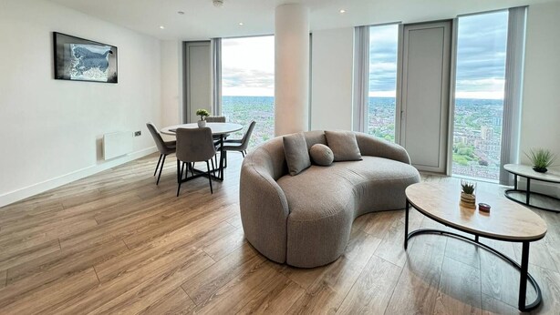 Una sala de estar moderna con un sofá curvo, una mesa de centro redonda y un comedor con sillas. Amplias ventanas ofrecen vista a la ciudad.