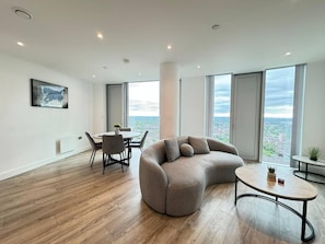 A modern living room with a curved sofa, a round coffee table, and a dining area with chairs. Large windows offer a city view. - Luxurious Deansgate 3bedroom Apartment Sleeps 10 (Manchester)
